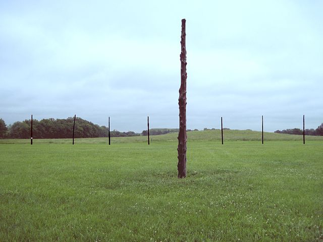 Wooden posts spaced in equal distance from each other in a circle on a green field. In the center is one post.